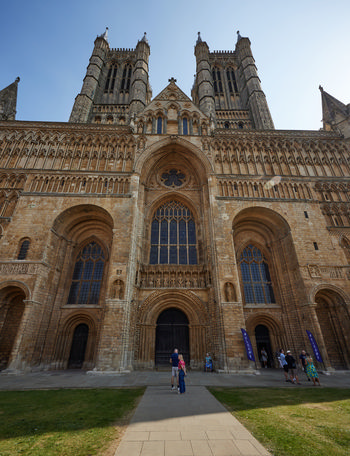 Lincoln Cathedral entrance This architectural photograph shows the grand entrance of Lincoln Cathedral, a prominent church located in the city of Lincoln, England, United Kingdom. The image captures the impressive façade of the cathedral with its towering spires and intricate stonework characteristic of English medieval architecture. Taken during the late morning in early autumn, the photograph features clear skies and sunlight casting shadows across the entrance and surrounding area. Lincoln Cathedral, a significant landmark in the region, stands out with its detailed gothic style and historical significance as a major cathedral in England. Visitors can be seen near the doorway, contributing a sense of scale to the imposing structure. The overall scene highlights the architectural beauty and cultural importance of Lincoln Cathedral within the United Kingdom.